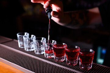 A bartender is pouring bright red liqueur into a line of shot glasses on the bar counter. Half of the glasses are already filled, creating a vivid visual contrast between the empty and full ones