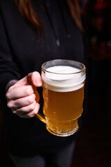 A person in a black hoodie holding a mug of light beer. The drink has a thick foam head, captured in a bar-like setting with focus on the glass mug and freshness of the beverage