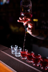 A bartender is pouring bright red liqueur into a line of shot glasses on the bar counter. Half of the glasses are already filled, creating a vivid visual contrast between the empty and full ones