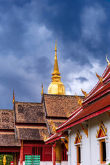 golden chedi and ornate rooftops of wat phra singh in chiang mai stand out beneath dramatic storm...