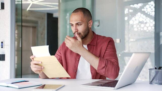 Upset disappointed businessman reading a letter with bad news sitting in a business office. Worried entrepreneur frustrated by receiving unpleasant negative notification after opening the envelope