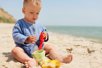 A curious baby in a blue outfit plays with sand and colorful beach toys on a sunny beach, with the sea in the background.