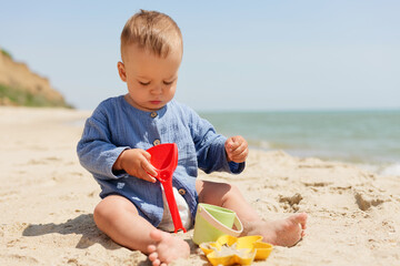 A cute baby in a blue outfit sits on a sunny beach, intently playing with a red spade and sand...