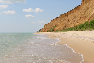 A sandy beach stretches along the coast with gentle waves and a large sandy cliff under a bright,...