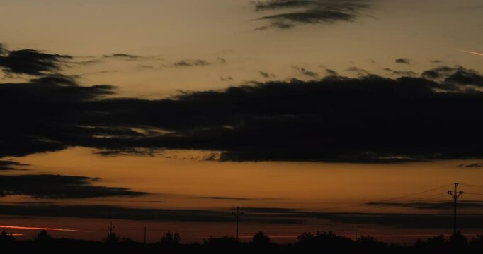 Dark Silhouette of a Low Horizon with Trees Bushes and Power Poles Against a Sky Lighting from Orange to Crimson Just Before Sunrise with Strips of Fast Moving Low Clouds Streaming Away from Camera