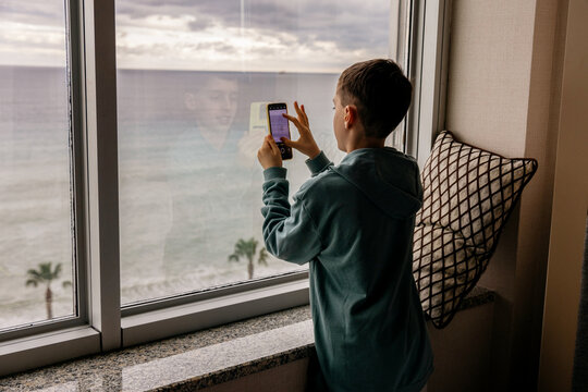 Boy with smartphone gazing out the window at sea view - Powered by Adobe