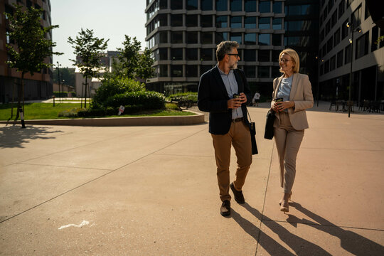Happy mature office colleagues in stylish outfits walking and talking on city street during summer