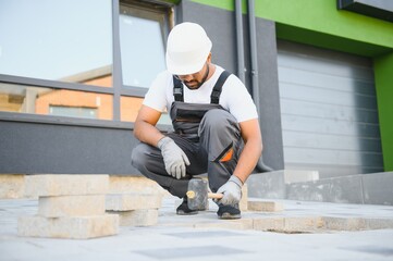 Indian master in gloves lays paving stones in layers. Garden brick pathway paving by professional paver worker. Laying gray concrete paving slabs in house courtyard on sand foundation base