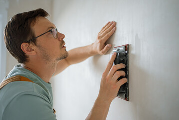 Construction worker carefully sanding a white wall with sandpaper during a home renovation project, ensuring a smooth and even surface for painting or wallpapering © wifesun