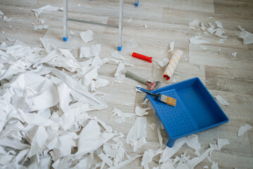 Paint tray, brush, roller, scraper, and numerous wallpaper scraps scattered across the floor,...