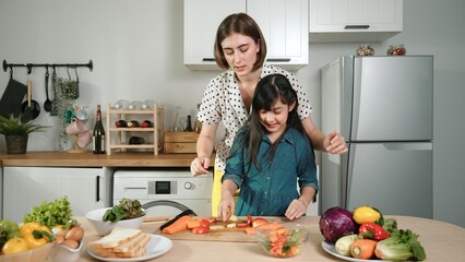 Smart caucasian mother and asian girl cooking together and chopping vegetable or preparing salad for dinner. Happy mom and daughter making healthy food with fresh food. Healthy food concept. Pedagogy.