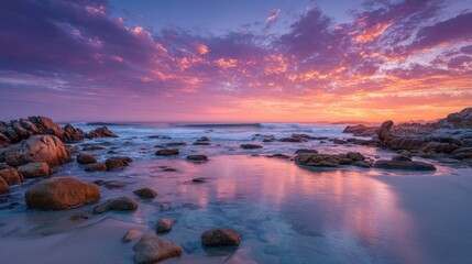 Colorful clouds reflect on the water as the sun sets over a serene beach. Rocks line the shore, creating a peaceful atmosphere in the twilight.