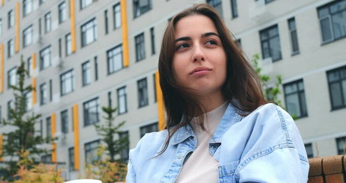 Stylish young woman sits on a bench, savoring a hot beverage from a disposable cup, taking a break amidst the backdrop of a modern building
