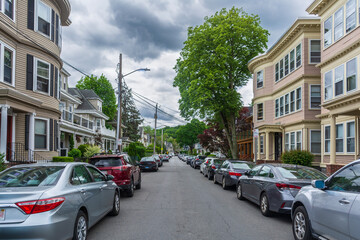 Fototapeta premium Multi-story homes lining a narrow road with parked vehicles and trees in Brighton, Massachusetts, USA 