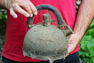 one closed lid with plastic black handle modern stainless steel dirty in cobwebs used discarded round empty teapot in hands of man in red t-shirt daytime near green leaves