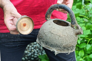 one open with lid and plastic black handle modern with stainless steel dirty in cobwebs used discarded round empty teapot in hands of man in red t-shirt daytime near green leaves