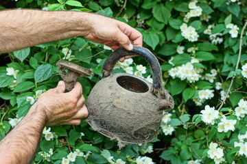 one open with lid and plastic black handle modern with stainless steel dirty in cobwebs used discarded round empty teapot in hands of man daytime near green leaves outdoors