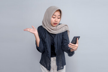 Asian Muslim woman in hijab looking confused while holding smartphone and raising one hand. She...