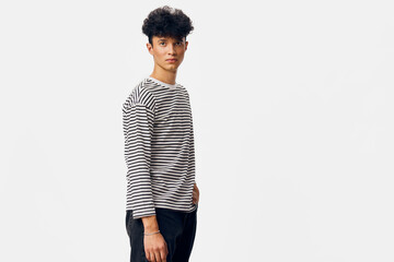 young man with curly hair wearing striped shirt and dark jeans stands confidently against white...