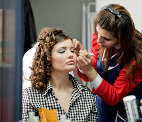 Makeup artist applying eyeliner to a young woman with styled curly hair in a beauty salon. Professional makeup session capturing elegance and transformation