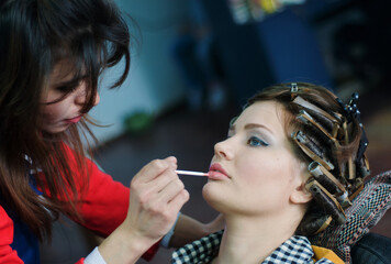 Makeup artist applying lip gloss to a young woman with hair curlers. Professional beauty preparation in a salon, highlighting glamour and personal care.