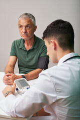 A man sits at a table in a clinic while a healthcare professional checks his blood pressure. The professional uses a digital monitor for the assessment.