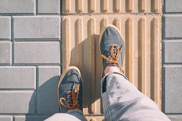 Women's feet in sneakers stand on a tactile tile. The road for the blind.	