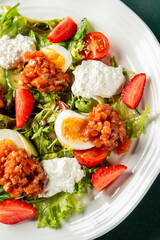 Fresh salad with boiled eggs, tomatoes, and strawberries served with creamy dressing on a white plate. Vibrant, healthy dish ideal for lunch or dinner menus.