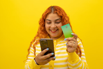 Cheerful young woman with curly red hair holding a card while shopping from her cell phone on a yellow background, concept of joy when shopping from cell phone