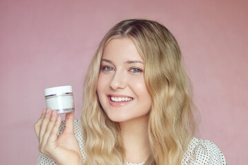 Beautiful blonde woman holding face cream jar in hand, smiling against soft pink background....