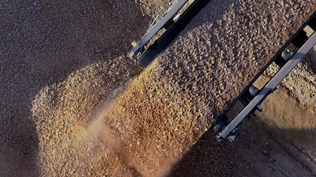 Top-down aerial shot following a mobile stone crusher's conveyor belt as it pours a steady stream of gravel, which is then moved and managed by a large wheel loader.
