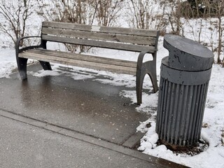 A Snowy Park Bench Alongside a Trash Can Set in a Beautiful Winter Landscape Scene. A serene snowy...