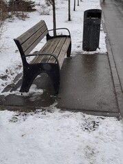 A Snowy Park Bench Alongside a Trash Can Set in a Beautiful Winter Landscape Scene. A serene snowy park scene depicting a wooden bench alongside a sleek, modern trash can, creating environment