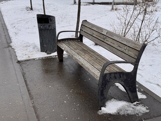 A Snowy Park Bench Alongside a Trash Can Set in a Beautiful Winter Landscape Scene. A serene snowy park scene depicting a wooden bench alongside a sleek, modern trash can, creating environment