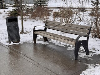 A Snowy Park Bench Alongside a Trash Can Set in a Beautiful Winter Landscape Scene. A serene snowy park scene depicting a wooden bench alongside a sleek, modern trash can, creating environment