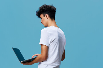 Young man using laptop with focus, standing against plain blue background, casual white t-shirt,...