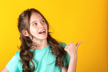 Portrait of cute young caucasian european blonde girl with opened mouth pointing thumb finger to the side side on yellow background. Studio shot