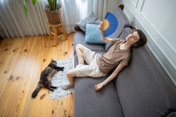 Exhausted woman fanning herself on sofa while cat lying nearby in heat distress. Indoor heat suffering, pet owner and animal under stress, summer heat without AC, discomfort, dehydration at home