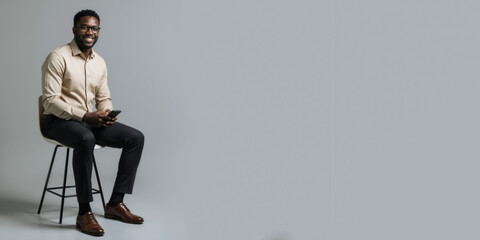 Young African American entrepreneur in beige shirt sitting on stool holding phone. Horizontal banner with confident happy black man holding smartphone against grey background with copy space for text.