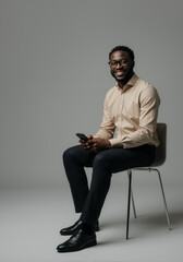 Young African American entrepreneur in beige shirt sitting on stool holding phone. Vertical portrait with confident happy black man holding smartphone against grey background