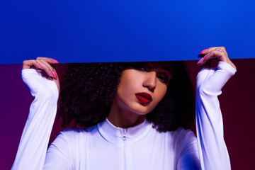 Trendy young model in a white bodysuit posing against vibrant neon purple and blue lighting,...