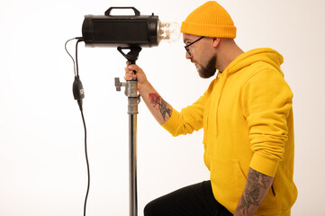 Smiling man in yellow hoodie holding professional lighting equipment in studio. Behind-the-scenes concept with cheerful crew member during photo or video shoot setup