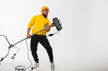 Smiling man in yellow hoodie holding professional lighting equipment in studio. Behind-the-scenes concept with cheerful crew member during photo or video shoot setup