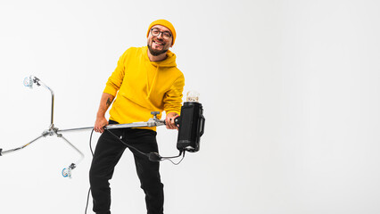 Smiling man in yellow hoodie holding professional lighting equipment in studio. Behind-the-scenes concept with cheerful crew member during photo or video shoot setup