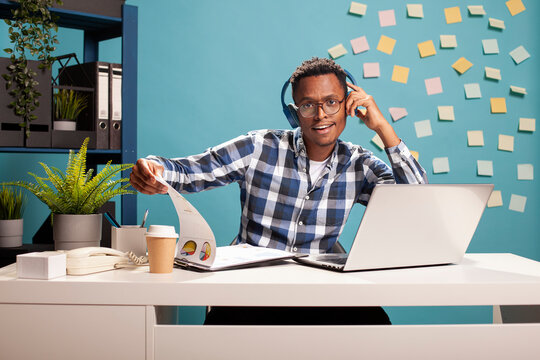 Joyful manager seated at desk, dancing and reviewing company information with paperwork and his laptop. Male analyst with headphones, flipping documents on clipboard and enjoying music on headphones.