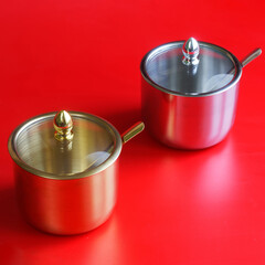 Two elegant metal sugar bowls, one gold and one silver, with glass lids and spoons on red...