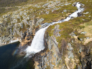 The power of Norway's wild nature: a drone view of a turbulent mountain river plunging from the cliffs as a powerful waterfall and creating a rainbow in the spray.
