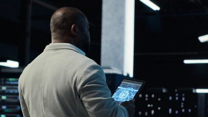 Server room software developer troubleshooting errors using software on laptop. African american IT professional walking in data center fixing bugs using notebook, camera B