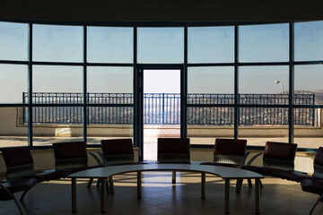 Modern waiting area with chairs and curved table in front of large glass windows and open door to terrace. Panoramic city view creates calm and spacious architectural atmosphere.