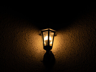Vintage outdoor wall lamp glowing at night on textured wall. Cozy warm light, home exterior, mood lighting concept